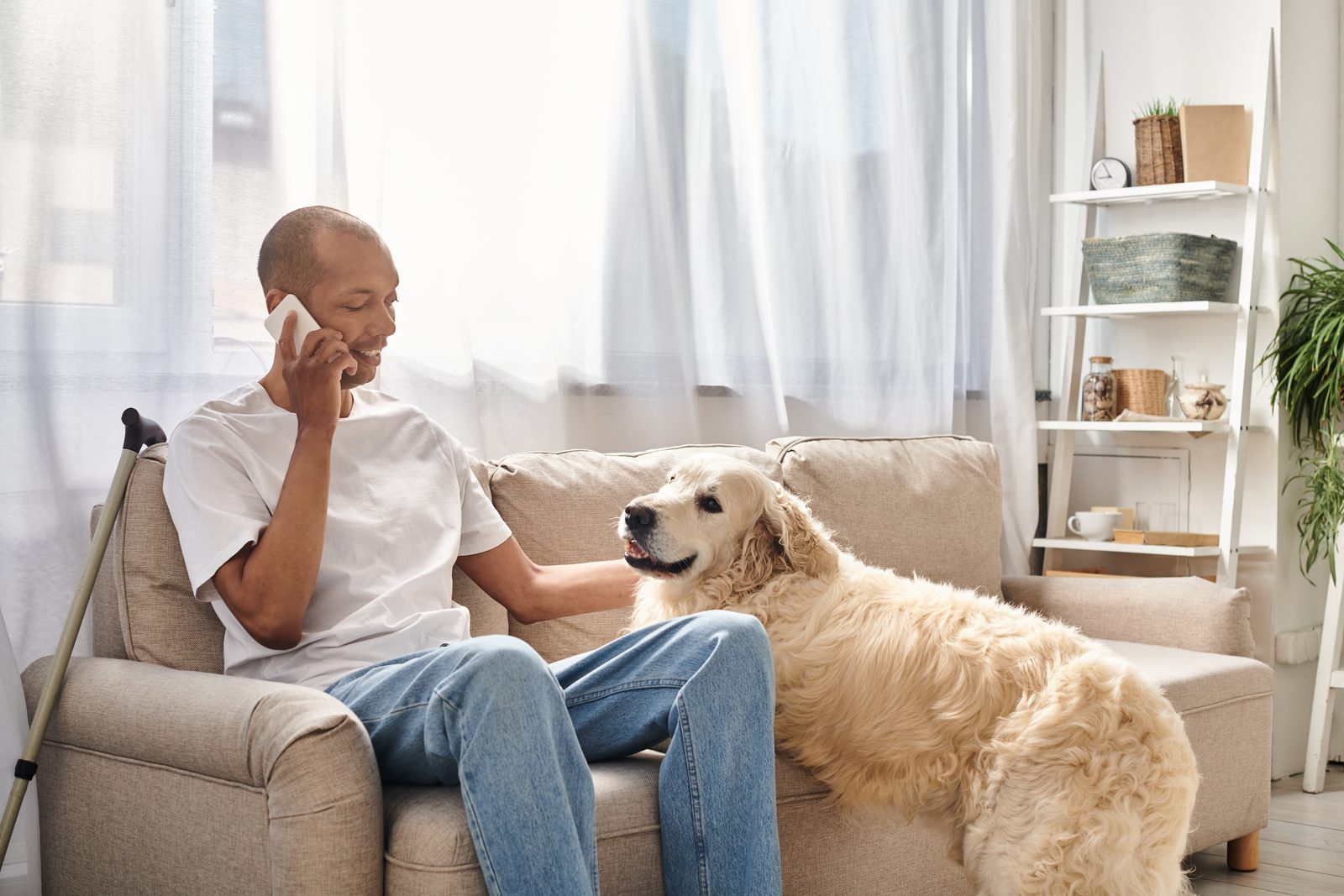 A man sits on a beige sofa talking on his phone while resting a hand on a golden retriever in a sunlit living room.