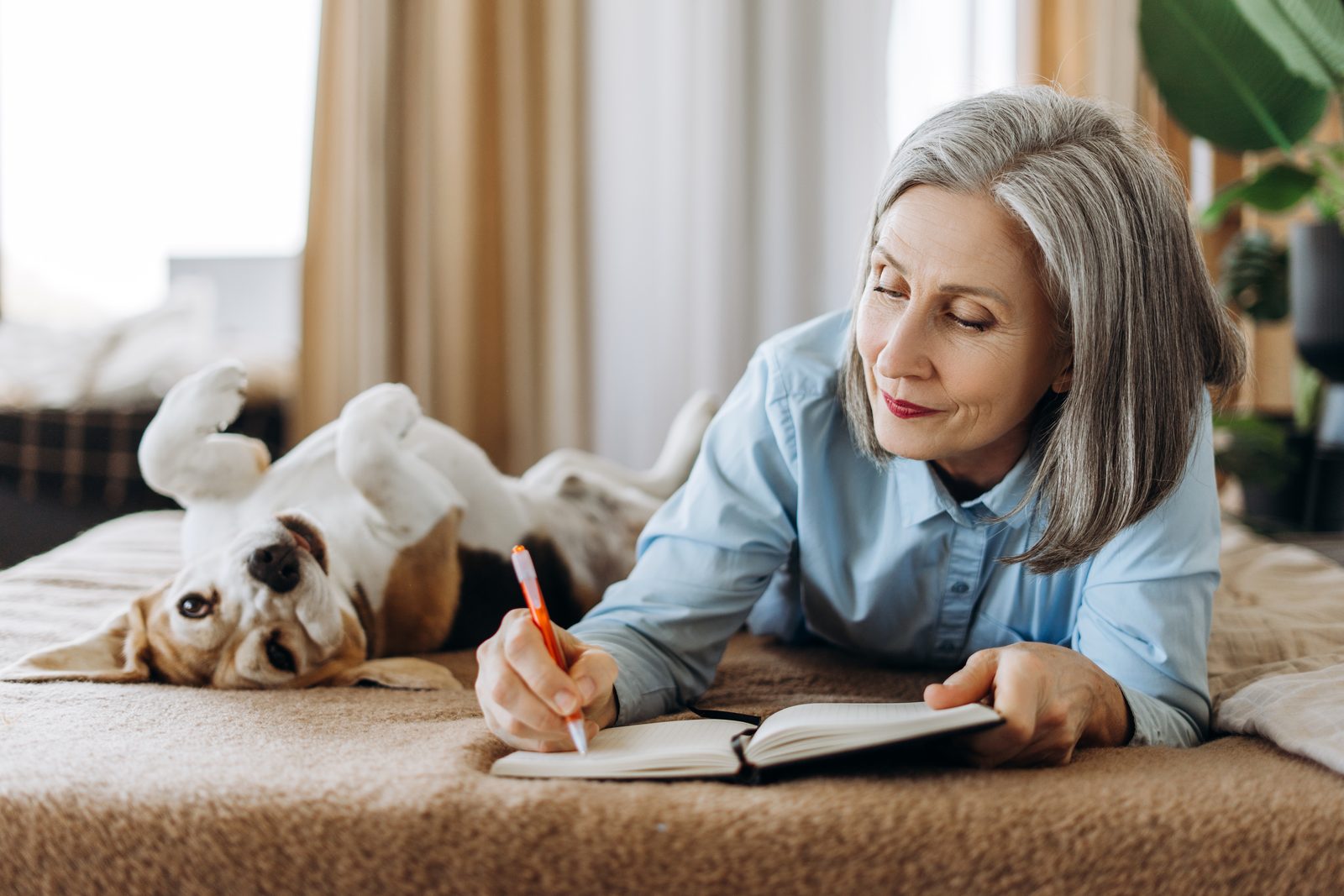 A gray haired woman lies on a bed writing in a notebook while a beagle rolls on its back beside her in a bright bedroom.
