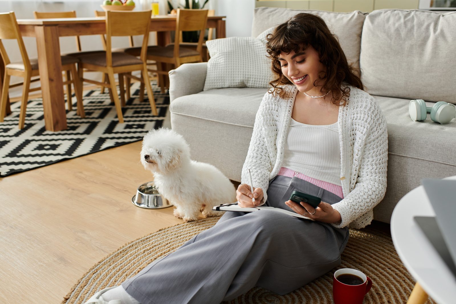 Woman sitting on a living room rug writing in a notebook while holding a phone, a small white fluffy dog sitting beside her
