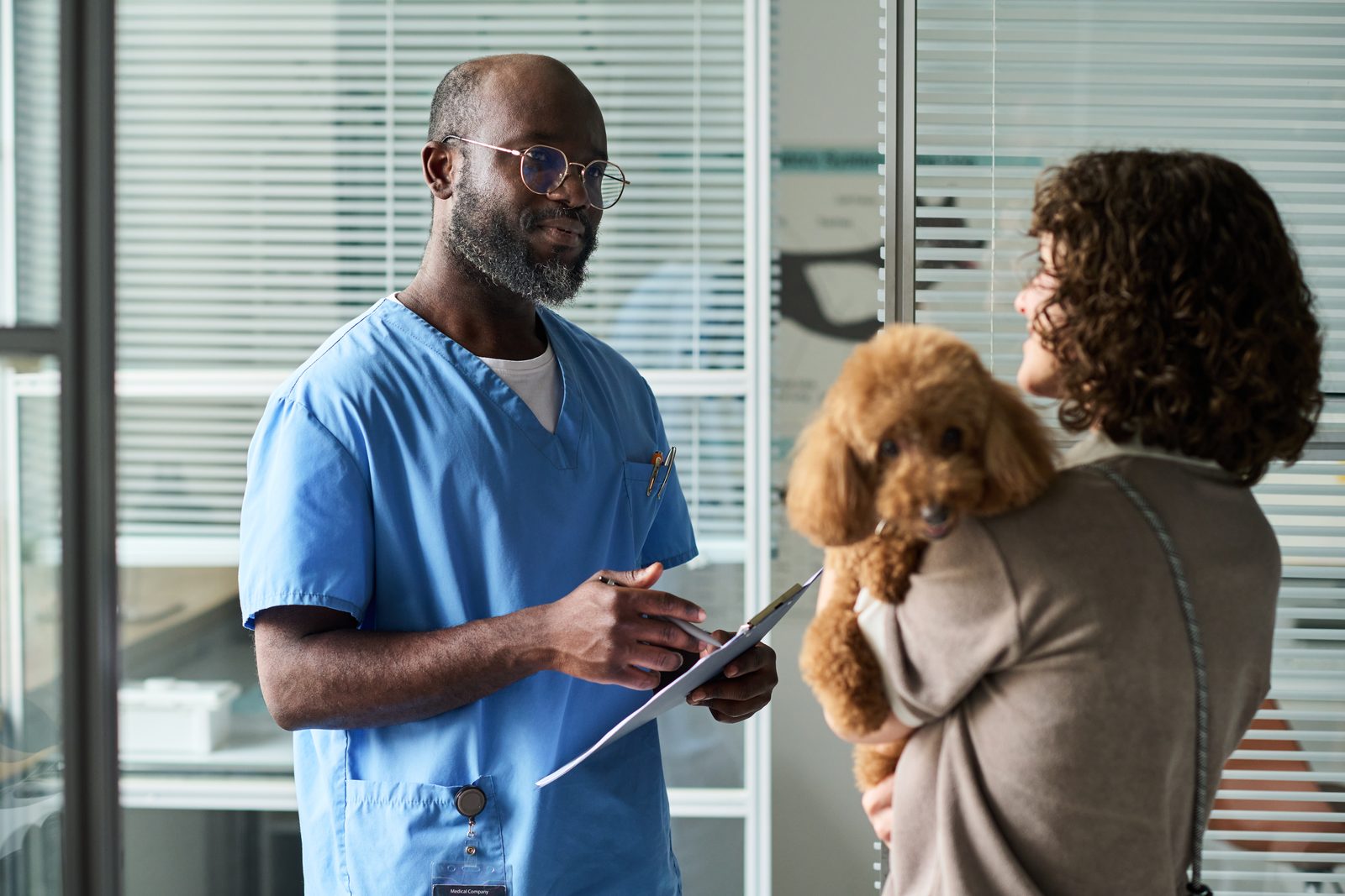 A veterinarian in blue scrubs writes on a clipboard while talking with a woman who holds a small curly-coated dog against her shoulder.