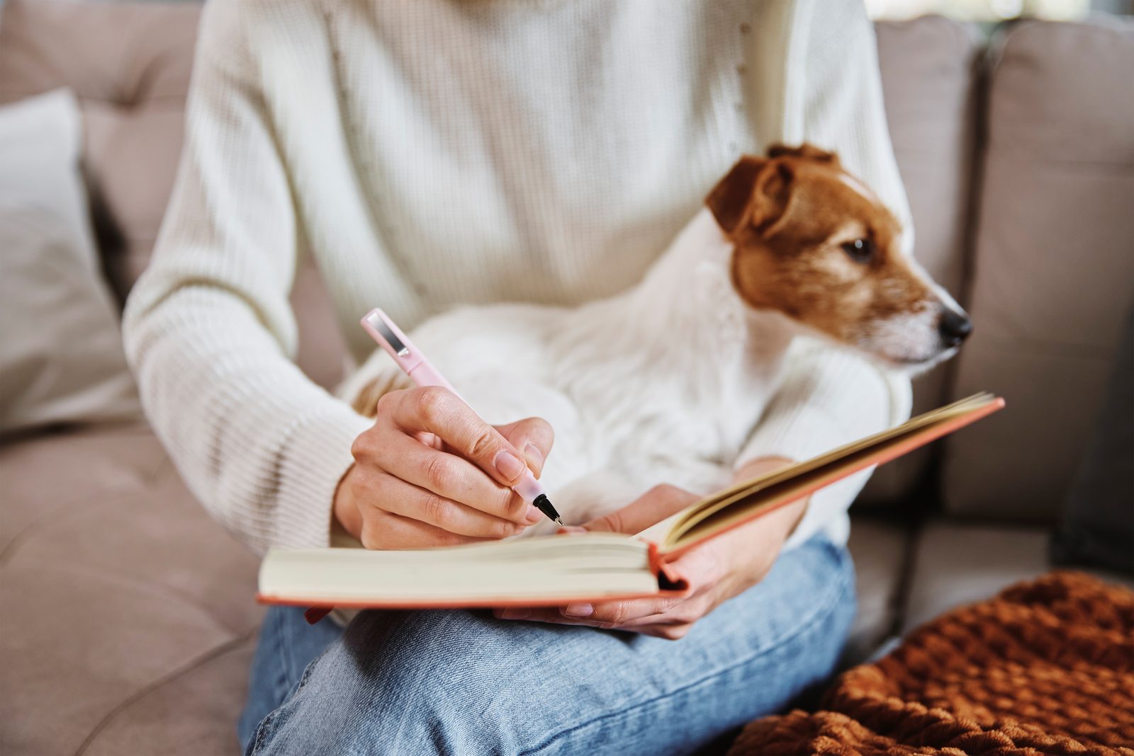 A woman in a cream sweater writes in an open notebook on the sofa while a Jack Russell terrier rests on her lap.
