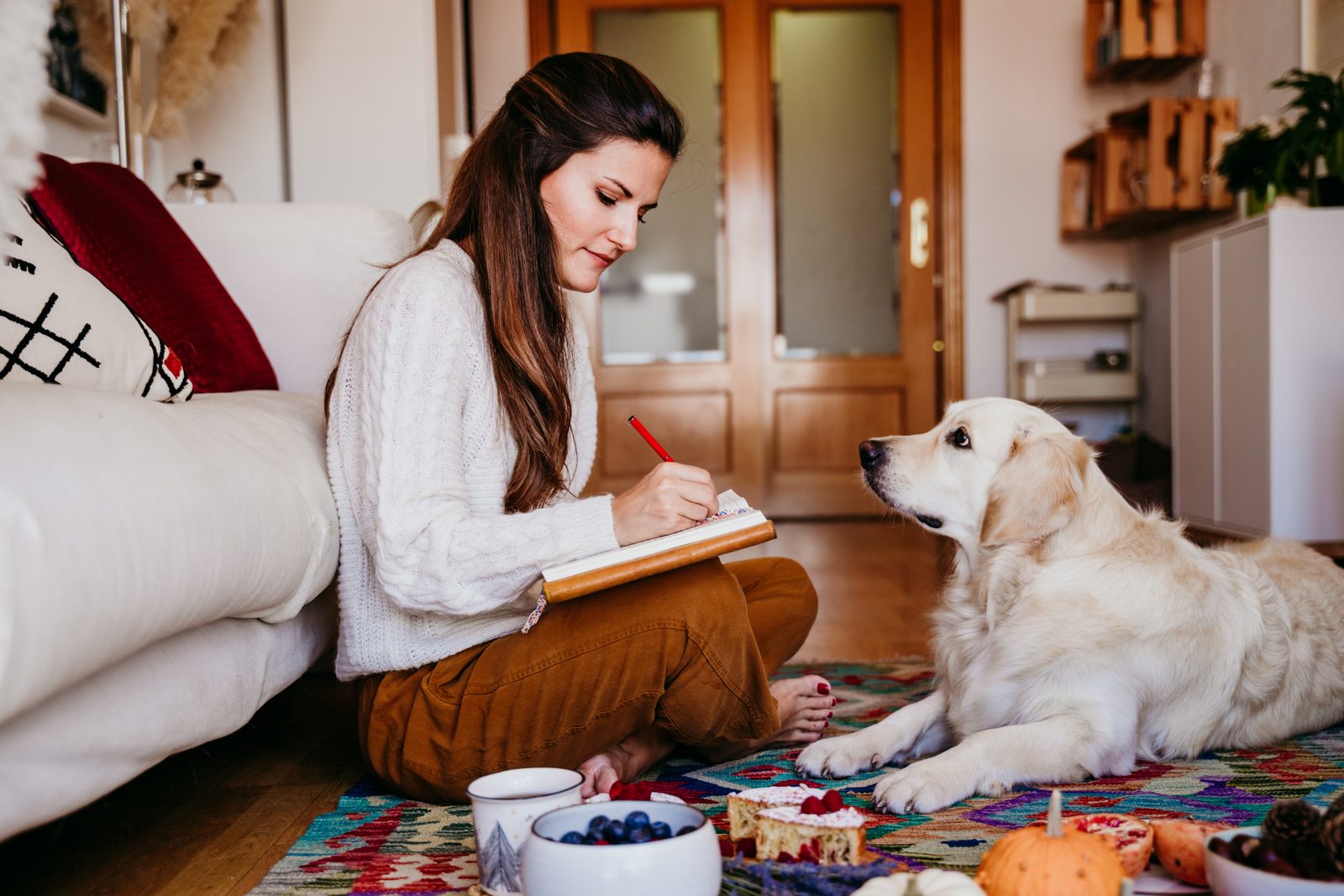 A woman sits on a living room rug writing in a notebook with a red pen while a golden retriever lies beside her, watching
